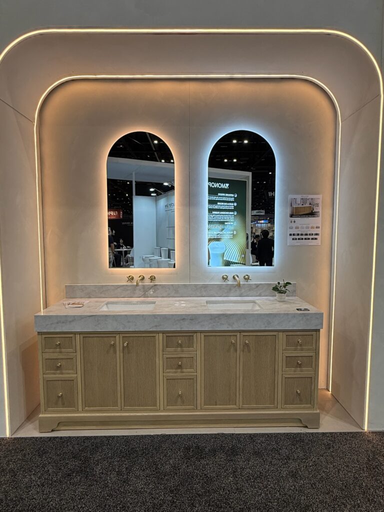 Wide light oak double sink fluted vanity with thick stone top. The vanity has 9 drawers, each with gold toned round knobs and two cabinets each with round knobs. Wall mounted gold toned faucets are above each sink. Two frameless back lit arched mirrors situated above each sink. 