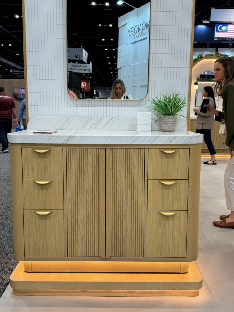 Rounded front corner vanity with white stone counter top.  The vanity is light oak wood, with brass pulls on each of its 6 drawers. The cabinet doors feature fluted wood on each of its two panels. There is a brass rounded rectangle mirror hanging above the vanity. 