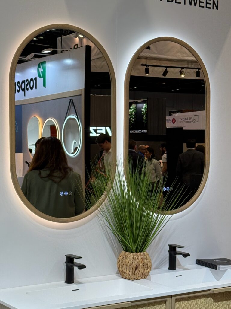 A set of oval bathroom vanity mirrors backlit with LED lighting and wood frames. They are above bronze faucets on a white stone vanity top. 
