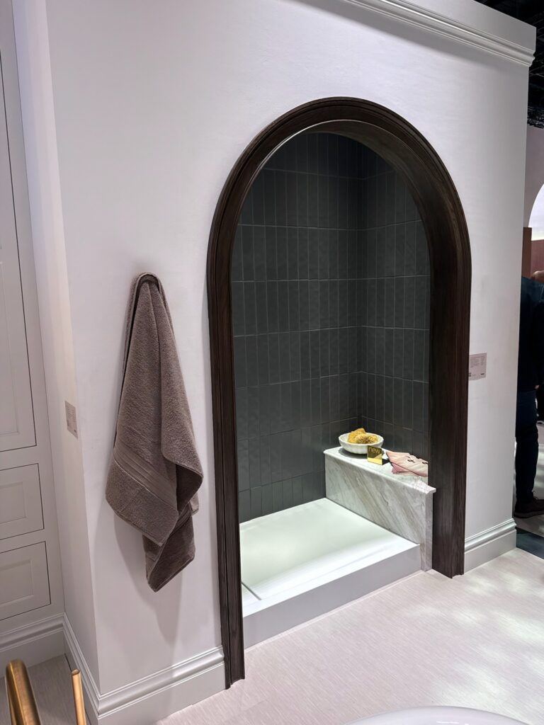 Arched Kohler shower alcove with dark wood trim. There is a stone shower bench with bathroom accessories on it.  There is a gray towel hanging to the left of the arched doorway. On the shower walls there is gray vertical tile. 