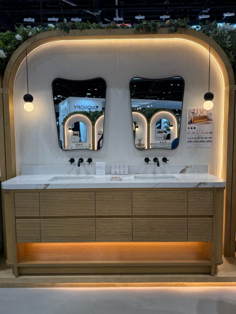 Long light oak vanity with horizontal fluting on the front of drawers.  There is a white stone top with light grey veining.  Two black  globe pendant lights hang to each side of each sink. Dual irregular shaped wall mounted mirrors with black fames are centered over each sink. Sleek wall mounted black faucets are situated below mirrors. 