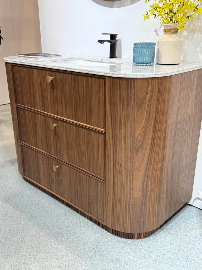 Contemporary bathroom vanity featuring three drawers, rounded front edges, and mahogany wood grain finish. White stone with grey veining and bronze single faucet. Off white vase with forsythia braces and blue glass candle to its left. 