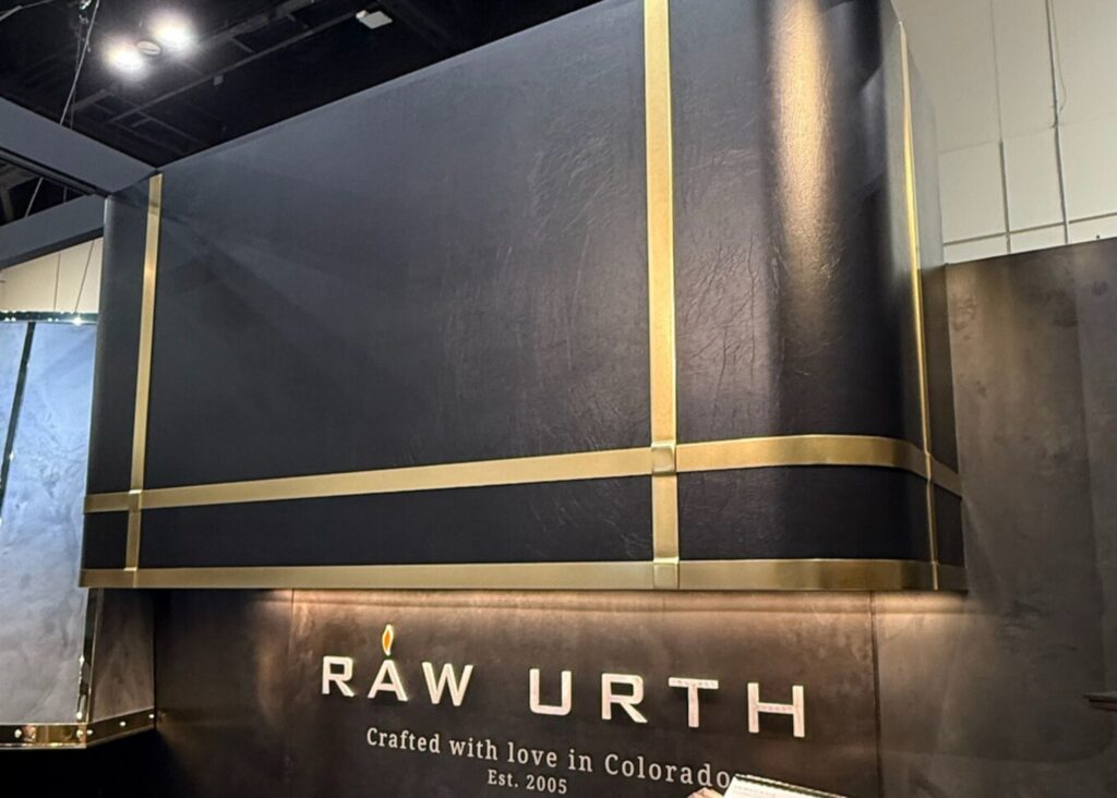 This image shows a large box shaped range hood that has rounded edges wrapped in textured black leather and decorative brass straps that add a modern industrial aesthetic. 